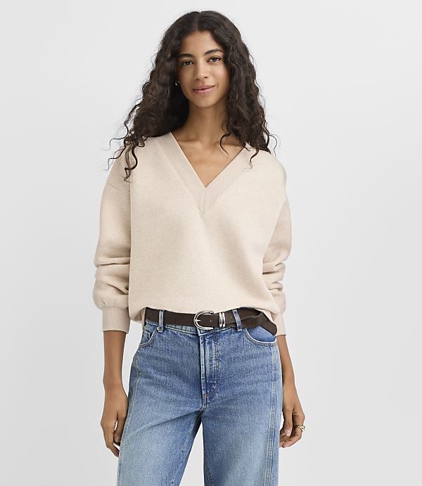A woman in a cream v-neck sweater and blue jeans stands against a plain white background.