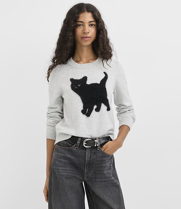 A woman in a light gray sweater with a black cat design stands against a plain white background.