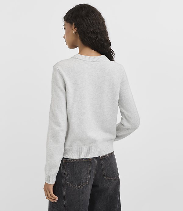 A woman in a light gray sweater and dark jeans stands against a plain gray background.