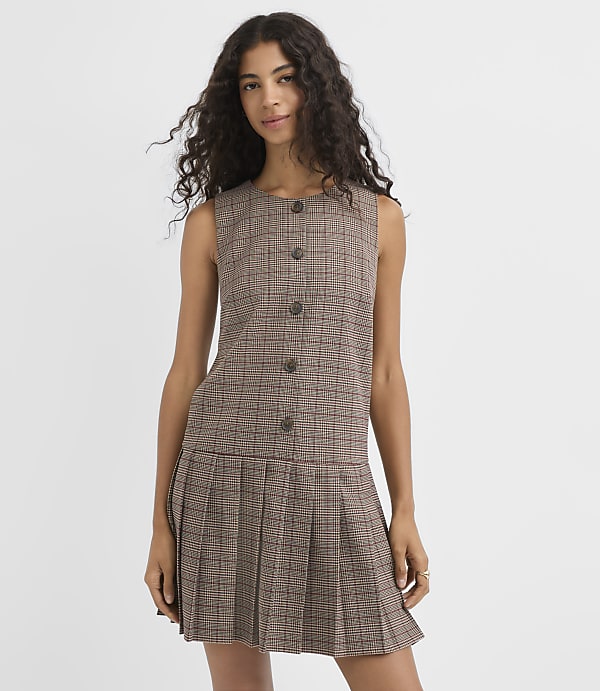 A woman with long, dark, curly hair stands in a sleeveless, plaid dress with a pleated skirt against a plain white background.