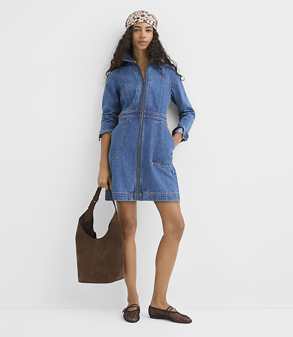 A woman in a denim dress and patterned headscarf stands against a plain white background, holding a brown suede bag and wearing brown flats.