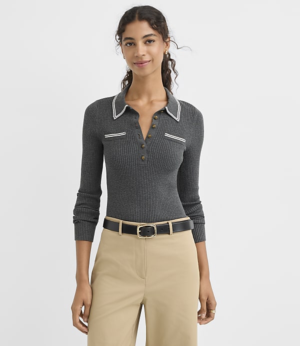 A woman in a gray ribbed polo shirt and tan pants stands against a plain white background.
