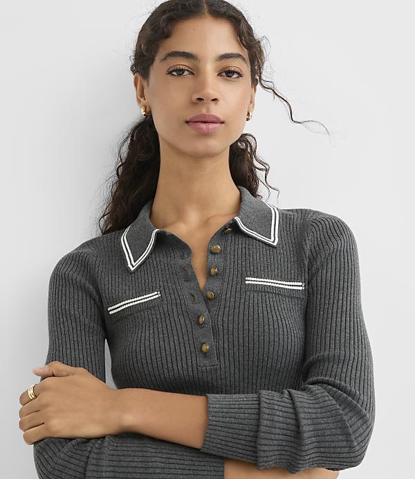 A woman in a dark grey ribbed polo shirt with white trim stands against a plain grey background.