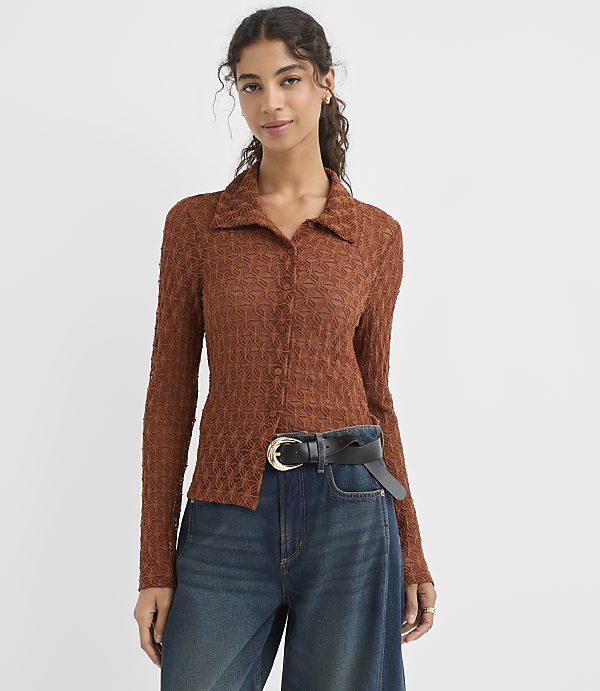 A woman in a textured brown collared shirt and blue jeans stands against a plain white background.