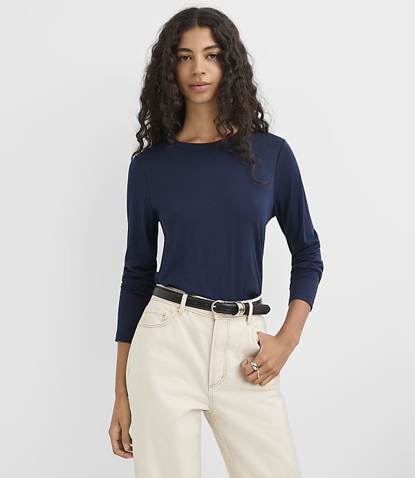 A woman in a navy long-sleeved shirt and cream-colored pants stands against a plain white background.