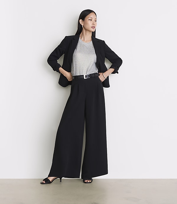 A woman in a black blazer and wide-leg pants stands against a plain white background.