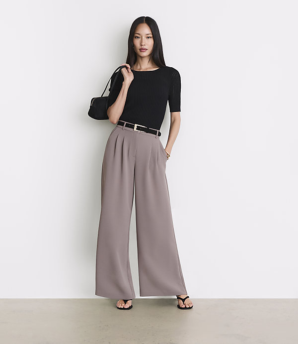 A woman in a black top and wide-leg pants stands against a plain white background.