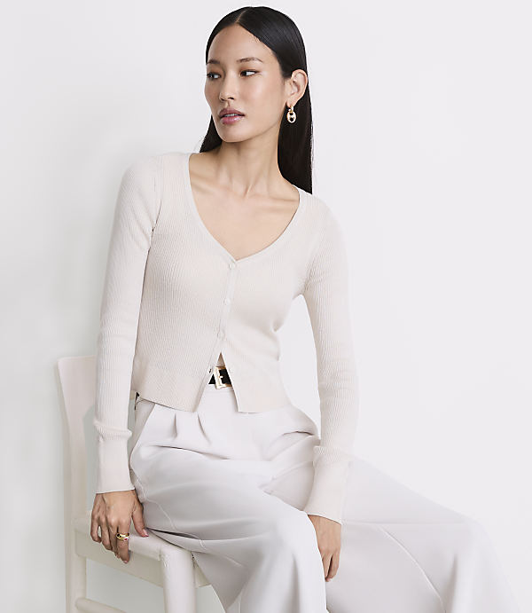 A woman in a cream cardigan and white pants sits on a white chair against a plain white background.