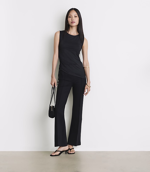 A woman in a black tank top and flared pants stands against a plain white background, holding a small black purse.