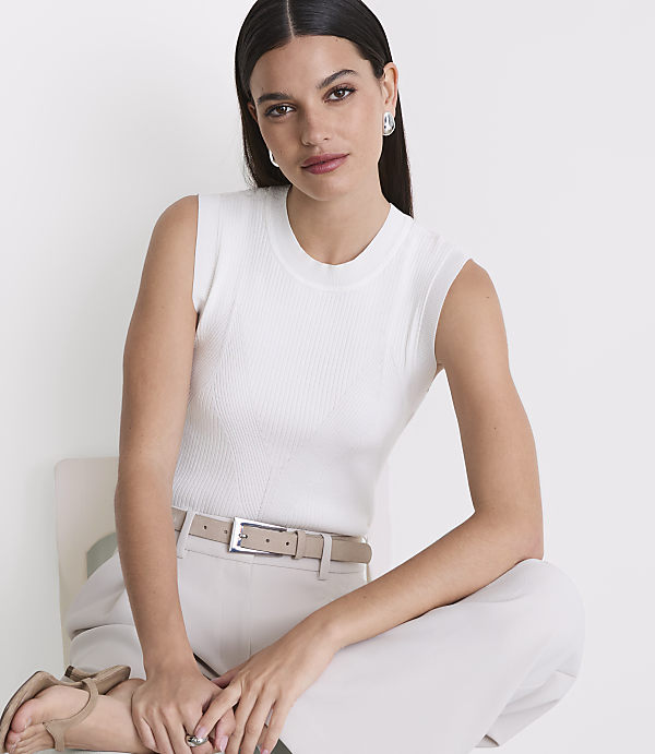A woman in a white ribbed tank top and beige pants sits on a chair against a plain white background.