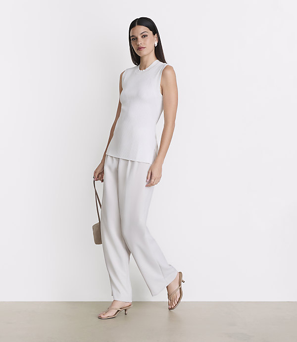A woman in a white sleeveless top and wide-leg pants stands against a plain white background, holding a small beige purse.