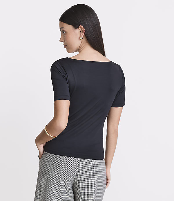 A woman in a black boatneck shirt and checkered pants stands against a plain white background.