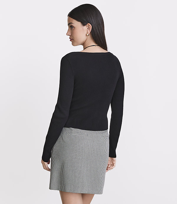 A woman in a black ribbed top and a checkered skirt stands against a plain white background.