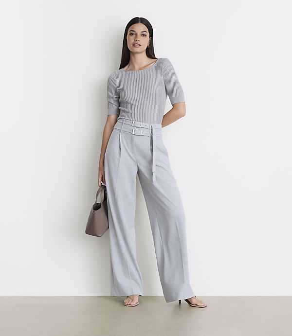 A woman in a grey ribbed top and light blue wide-leg pants stands against a plain white background, holding a taupe handbag.