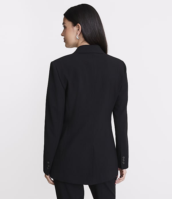 A woman in a black blazer stands against a plain white background.