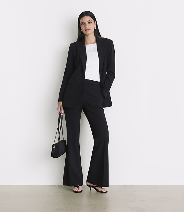 A woman in a black suit and white top stands against a white wall, holding a black purse.