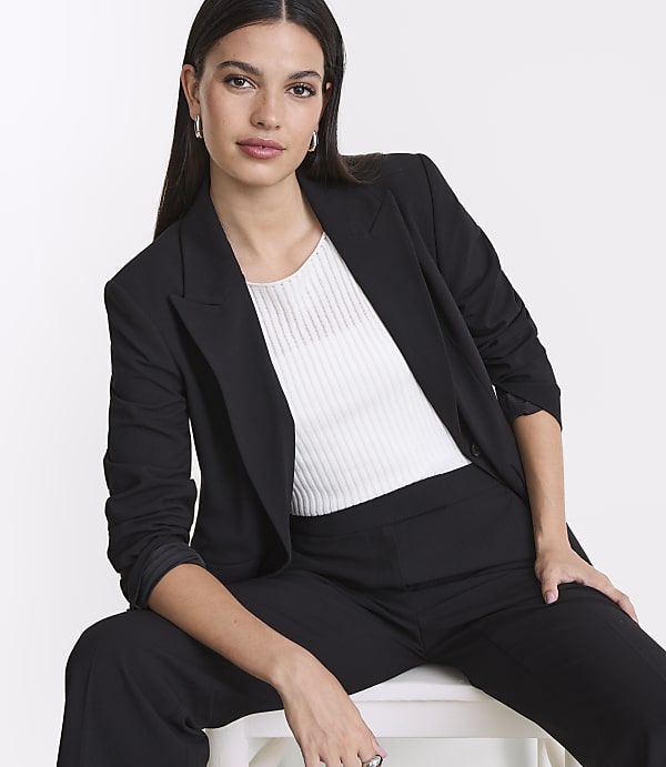 A woman in a black blazer and white top sits against a plain white background.