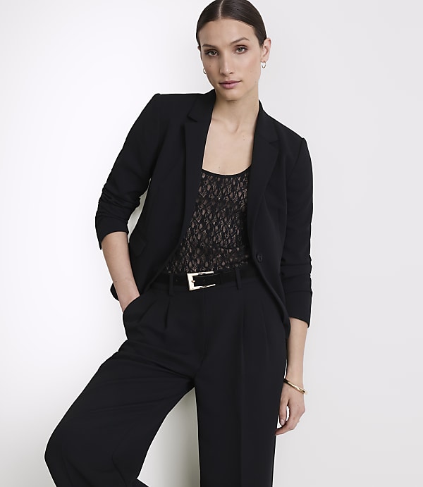 A woman in a black blazer and pants stands against a plain white background.