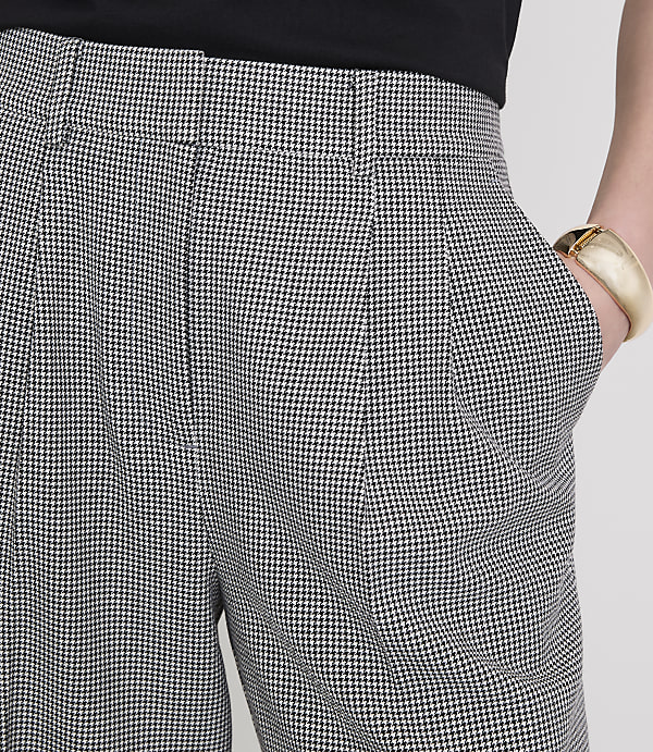 A person's hand, adorned with a gold bracelet, rests in the pocket of houndstooth patterned pants against a plain white background.