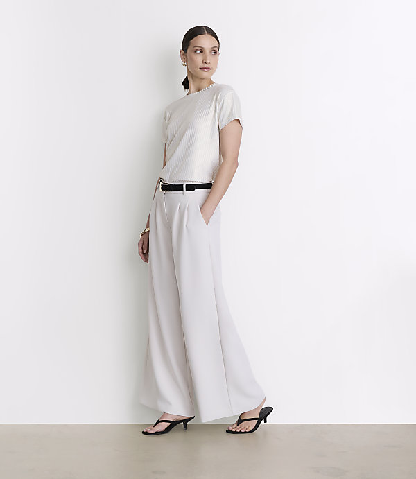 A woman in a white top and wide-leg pants stands against a plain white background.