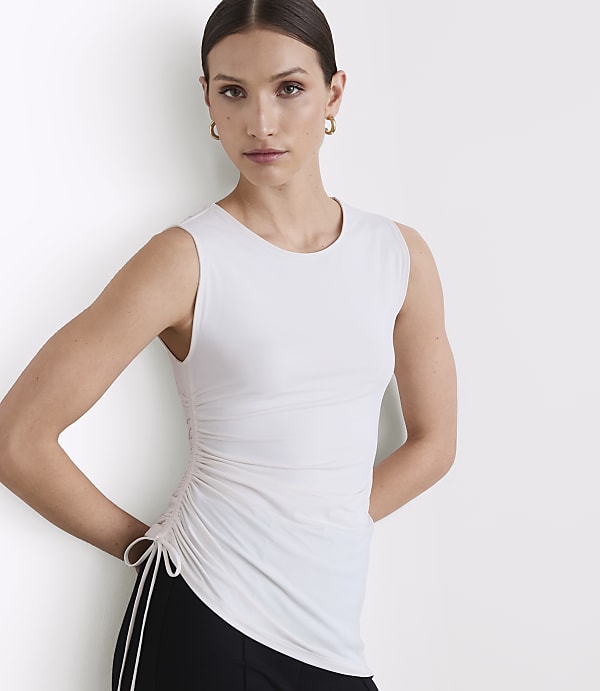 A woman in a white ruched tank top and black pants stands against a plain white background.