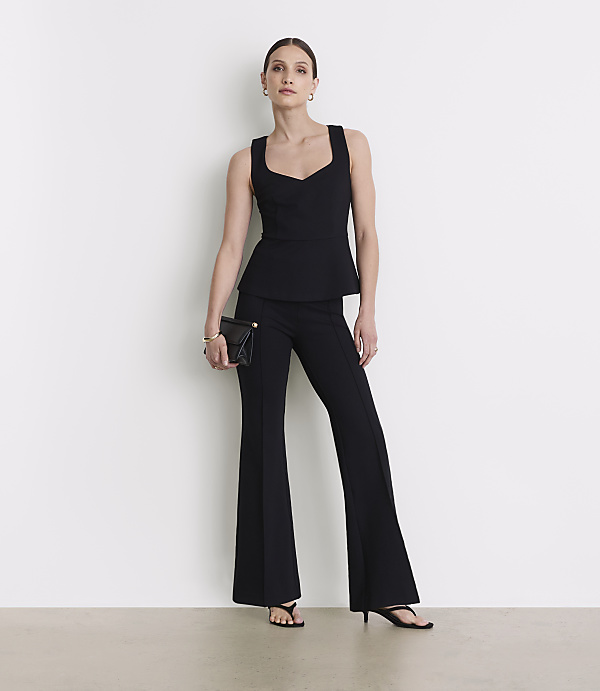 A woman in a black peplum top and flared pants stands against a plain white background, holding a black clutch.
