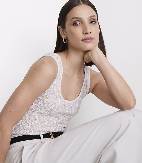 A woman in a white lace tank top and light-colored pants rests her chin on her hand against a plain white background.
