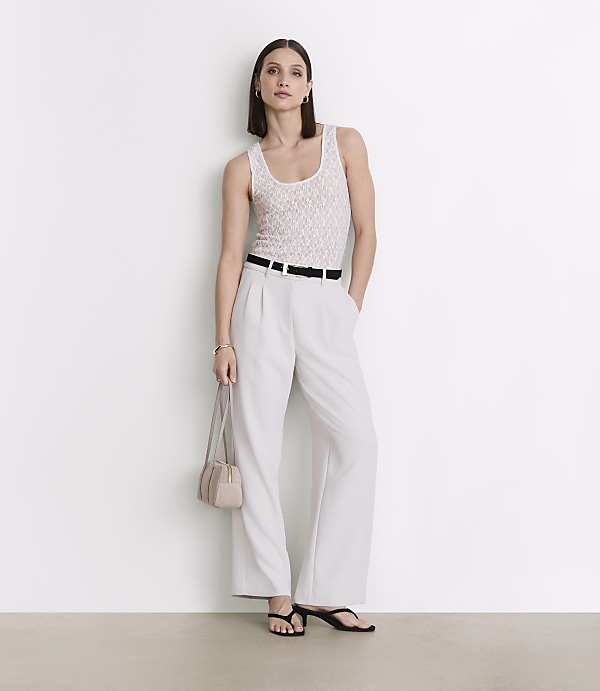 A woman in a white tank top and light-colored pants stands against a plain white wall, holding a small purse.