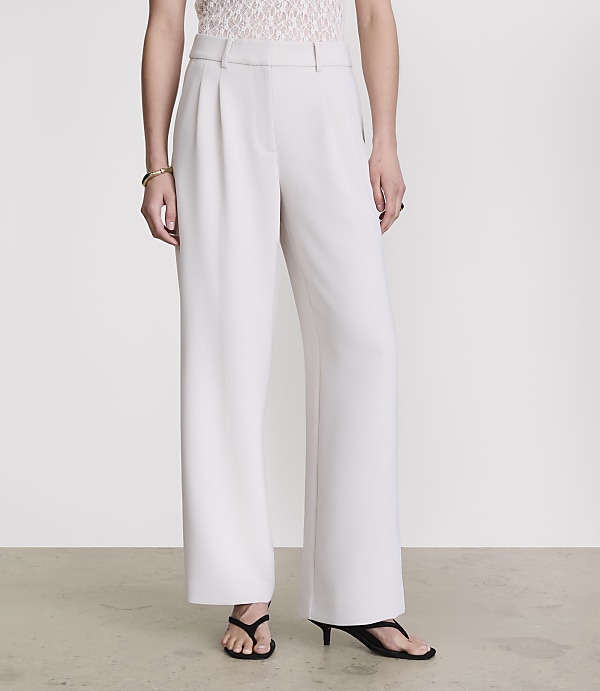 A person stands in white wide-leg trousers and a lace top against a plain grey background.