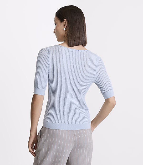 A woman in a light blue ribbed sweater and grey pinstripe pants stands against a plain white background.