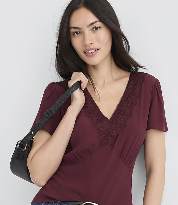 A woman in a burgundy top with lace trim holds a black purse in front of a plain white background.