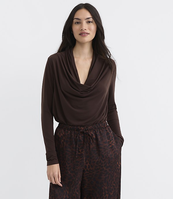 A woman in a brown cowl-neck top and leopard-print pants stands against a plain white background.