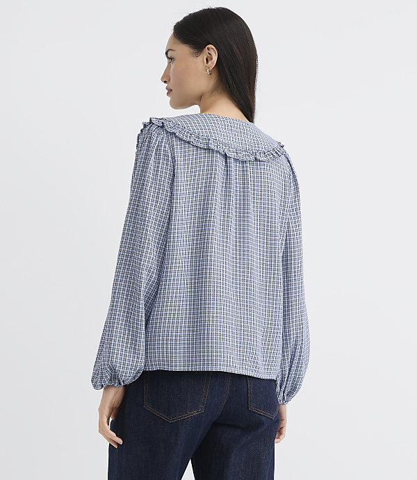 A woman in a blue and white checkered blouse with a ruffled collar stands against a plain white background.