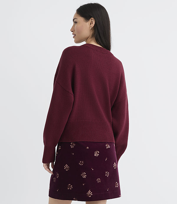 A woman in a maroon sweater and patterned skirt stands against a plain background.