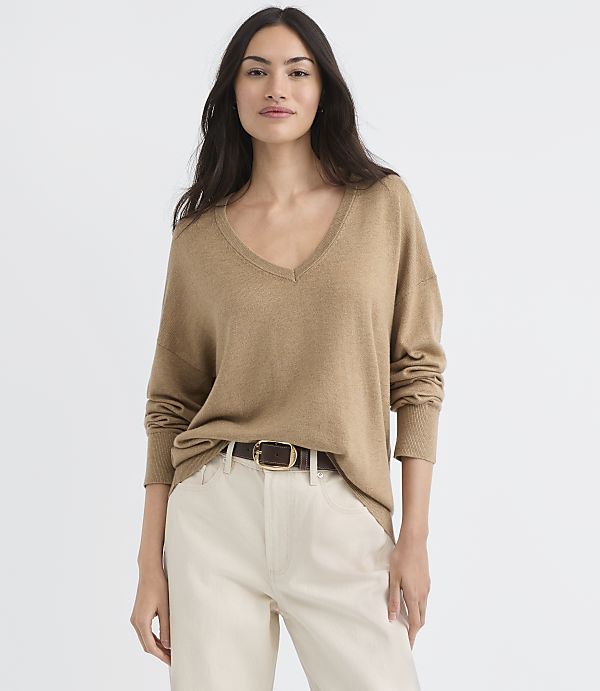 A woman in a tan v-neck sweater and cream pants stands against a plain white background.