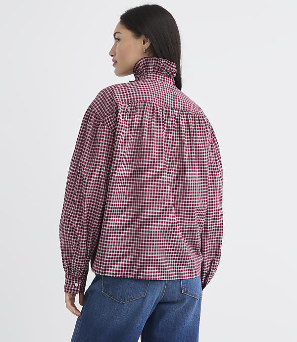 A woman in a red and black checkered blouse with a ruffled collar stands against a plain white background.