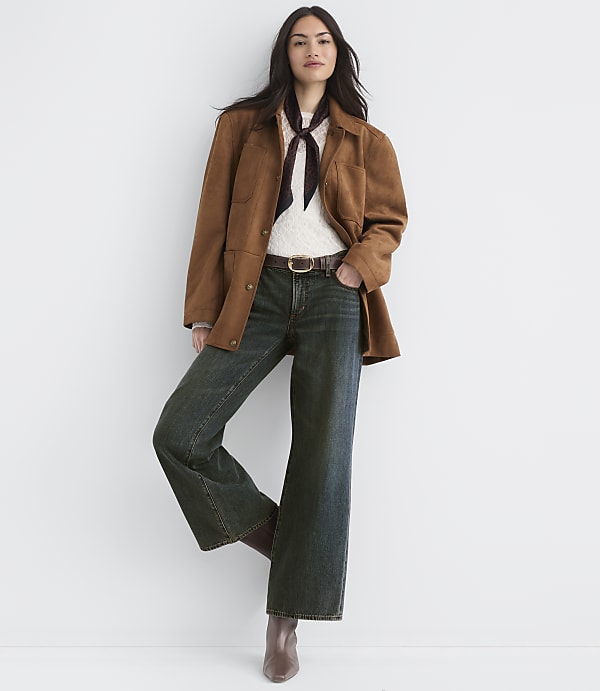 A woman in a brown jacket and wide-leg jeans stands against a plain white background.