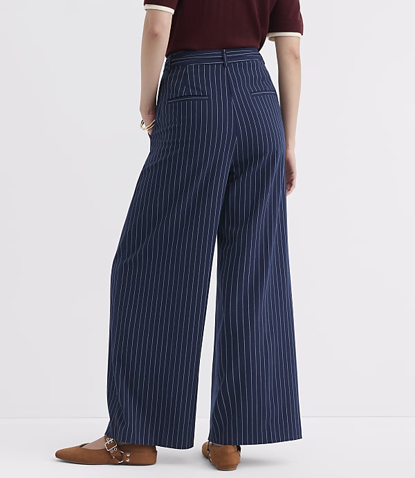 A person wearing navy pinstripe wide-leg pants and brown flats stands against a plain white background.