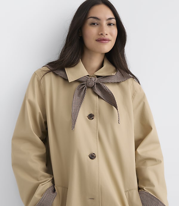 A woman in a tan trench coat with a striped collar and tie stands against a plain white background.