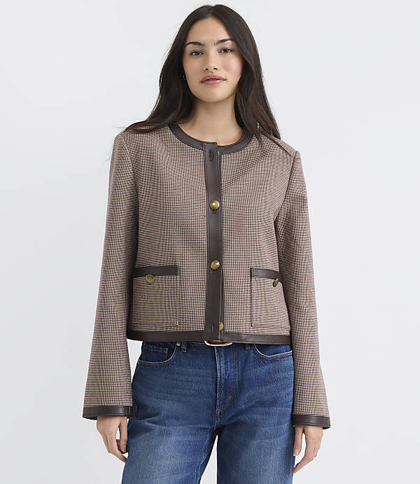 A woman in a brown patterned jacket with leather trim and blue jeans stands against a plain gray background.
