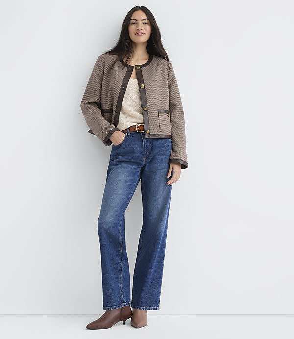A woman in a brown patterned jacket and blue jeans stands against a plain white background.