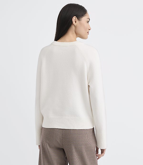 A woman in a cream sweater and plaid pants stands against a plain gray background.