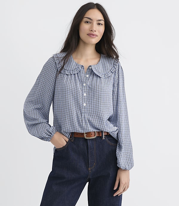 A woman in a blue checkered blouse and dark jeans stands against a plain white background.