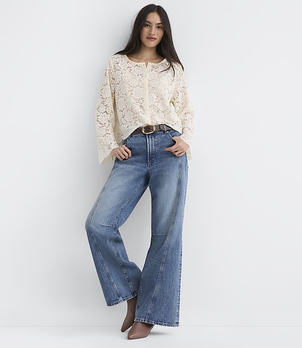 A woman in a cream lace top and wide-leg jeans stands against a plain white background.
