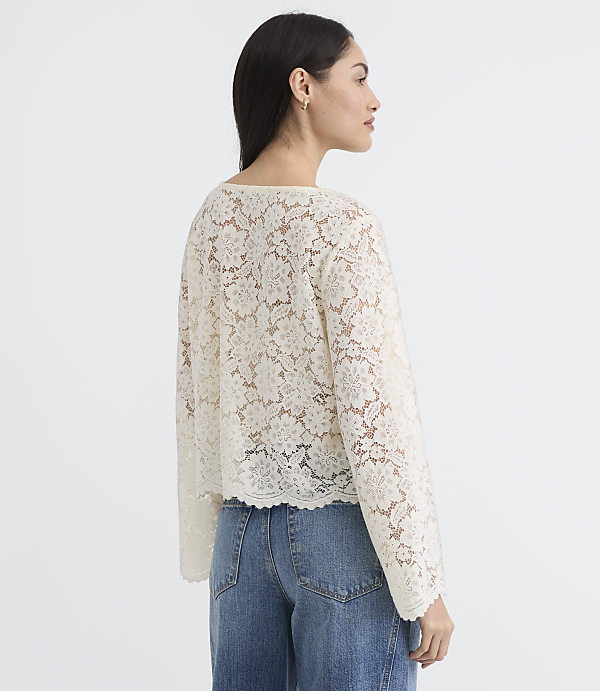 A woman in a cream lace top and blue jeans stands against a plain white background.