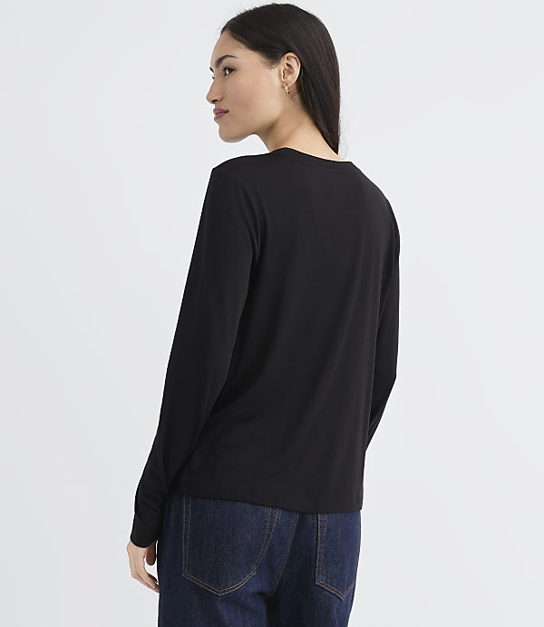 A woman in a black long-sleeved shirt and dark jeans stands against a plain white background.