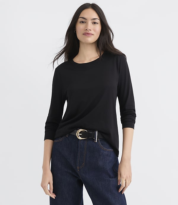 A woman in a black long-sleeved shirt and dark jeans stands against a plain white background.