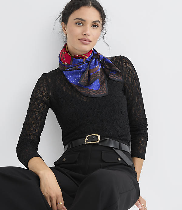 A woman in a black lace top and black pants with a patterned scarf tied around her neck sits against a plain white background.