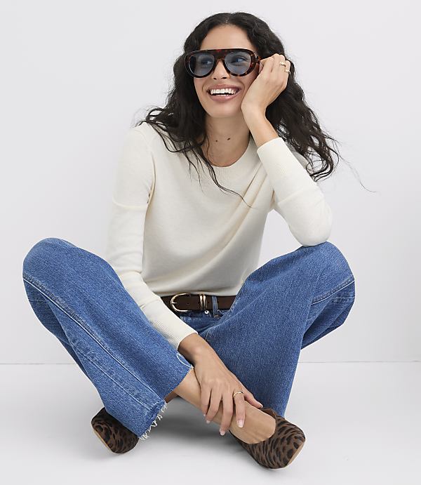 A woman in a cream sweater and wide-leg jeans sits cross-legged on a white background, wearing leopard print flats and sunglasses.