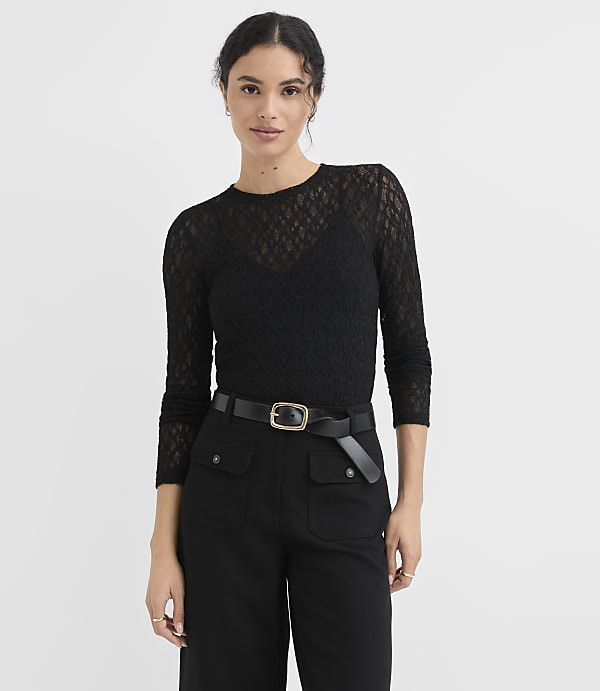 A woman in a black lace top and black pants with a gold-buckled belt stands against a plain white background.